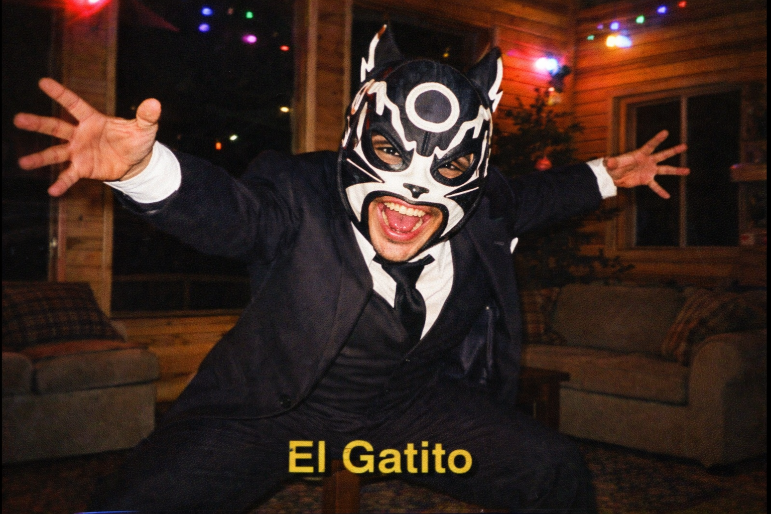 AI image prompt (360x240): A masked luchador in a crisp business suit doing an exaggerated wrestling entrance pose inside a cabin living room, colorful party lights, dramatic shadows, low-resolution 90s camcorder still frame, VHS scanlines.