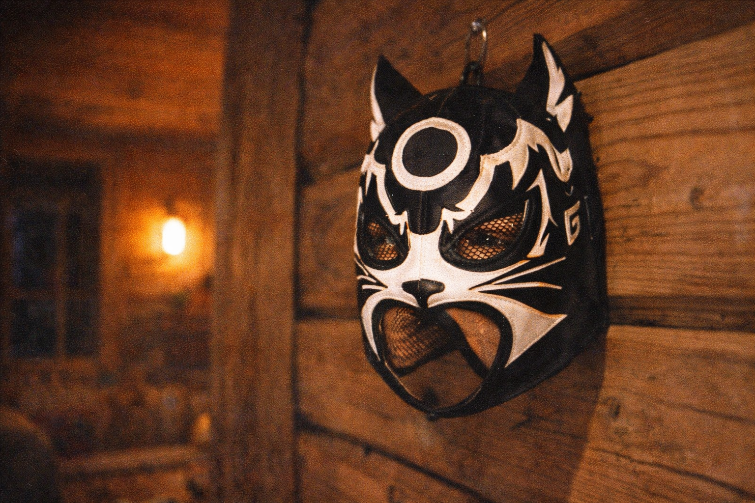 AI image prompt (360x240): Close-up of a black-and-white lucha mask with bold graphic shapes and mesh eye panels hanging on a wooden wall, warm cabin lighting, shallow depth of field, grainy 1999 photo.